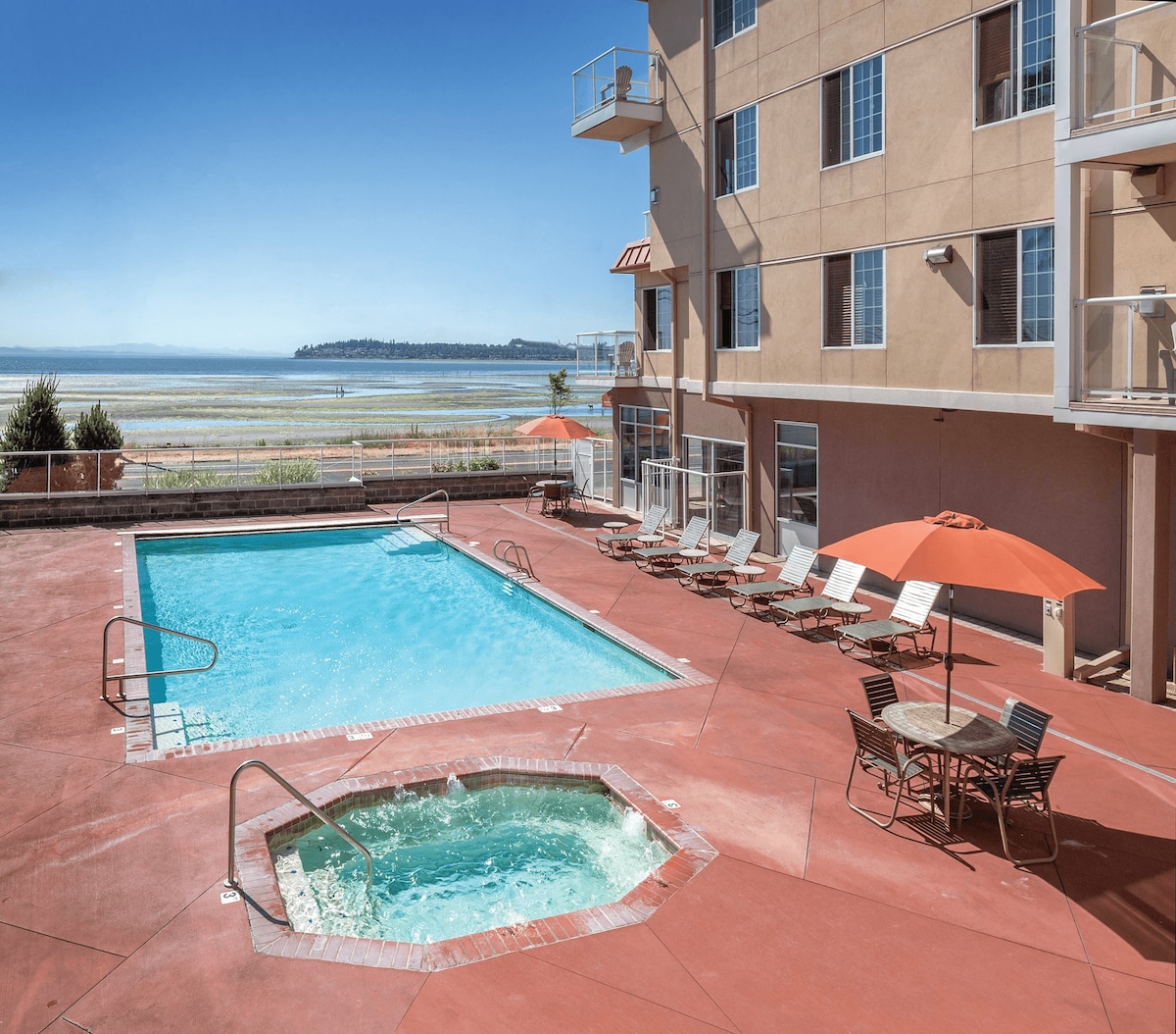 An outdoor pool is framed by a spacious patio area featuring lounge chairs and orange umbrellas. Adjacent to the pool, a hot tub is set within a tile border. The scenery includes a view of the water beyond the resort, under a clear blue sky.