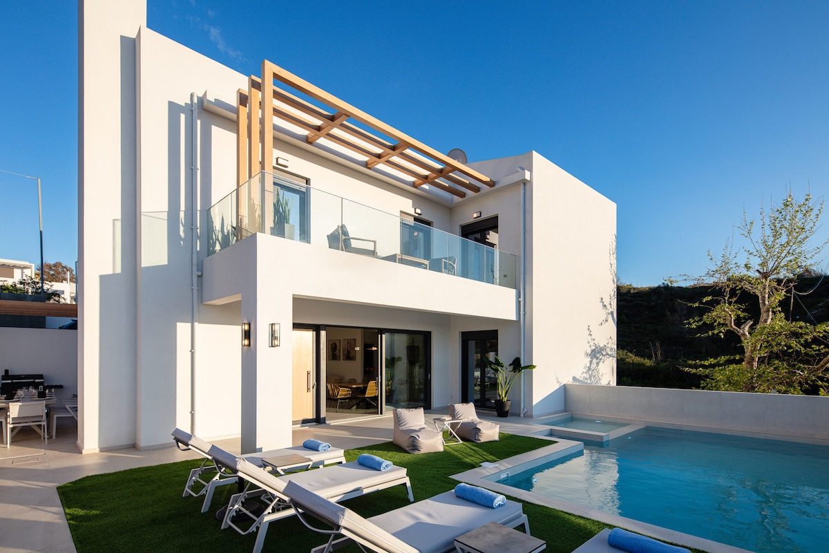 A modern house with a sleek design is highlighted against a clear blue sky. A spacious patio features several lounge chairs positioned near a calm pool, while large glass doors open to the interior. Lush greenery is visible in the background.