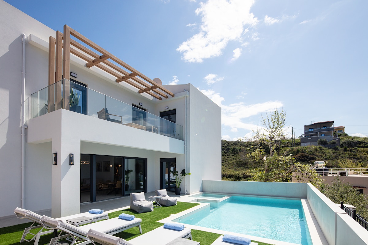 The exterior of a modern villa is displayed, featuring a light-colored facade with wooden accents. Lounge chairs are arranged around a clear blue swimming pool, while a balcony above offers views of the surrounding landscape under a bright sky.