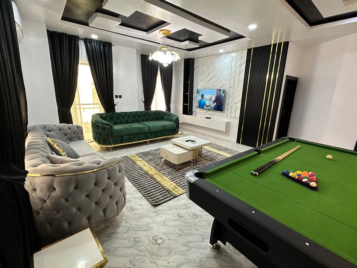 A stylish living area features a green velvet sofa and a tufted gray couch, complemented by a coffee table at the center. A game table with billiard equipment is present, while a television is mounted on the wall. Large windows are dressed with elegant black curtains.