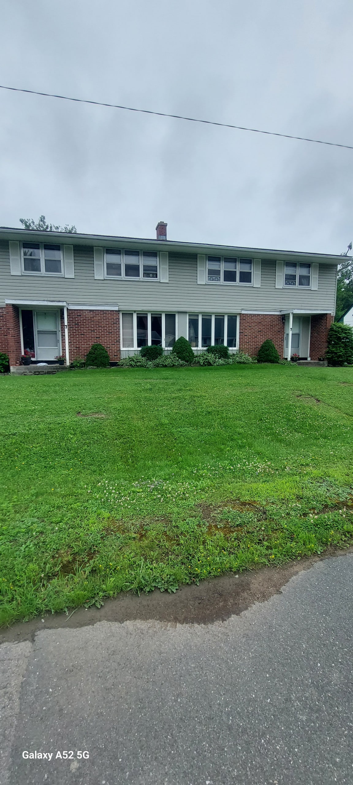 The exterior of a two-story house is presented, featuring a combination of green siding and brick. Large windows allow natural light into the rooms, while neat shrubs line the front. A well-maintained lawn provides an inviting entrance to the property.