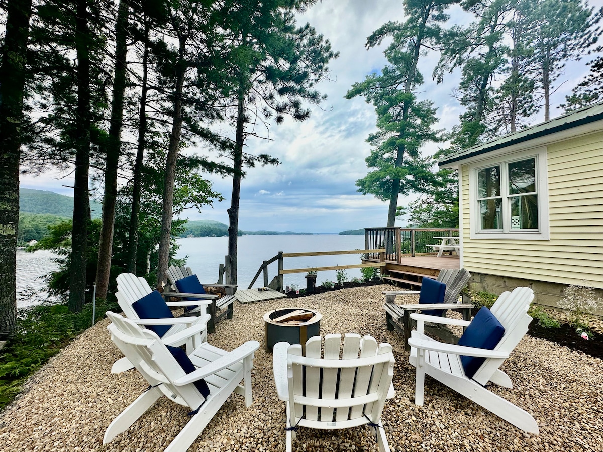 A quaint outdoor seating area features four white Adirondack chairs arranged around a central fire pit. The space is framed by trees, creating a serene view of the water and a nearby dock. A large deck is visible in the background.