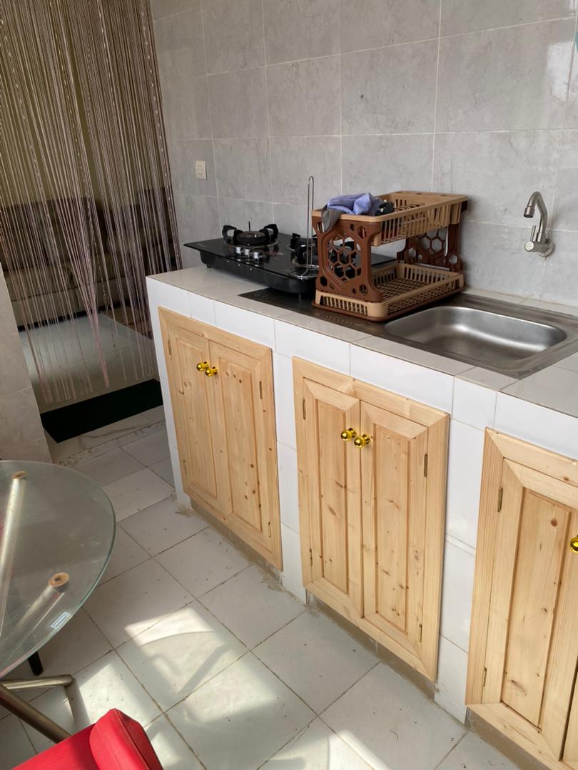 The kitchenette features light wooden cabinetry with gold handles and a stainless steel sink. A gas stove is positioned on the countertop, with a small wooden crate nearby. Natural light filters in, creating a bright workspace. A clear glass table and red chair are placed within view.