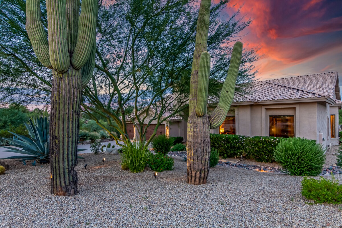 Large saguaro cacti rise prominently in a gravel landscape, surrounded by desert vegetation. A soft sunset casts warm hues across the sky, complementing the neutral tones of the home's exterior. Soft outdoor lighting accentuates the natural elements, creating an inviting outdoor space.