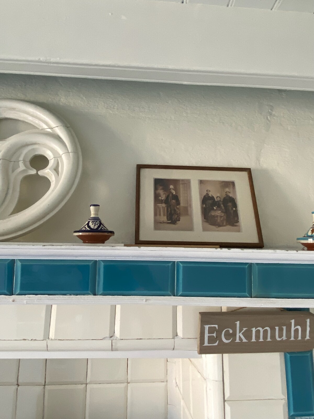 A decorative shelf is situated above a tiled surface, featuring a framed photograph of two figures in vintage attire. Small decorative items, including traditional pottery, are positioned on either side of the photograph. An ornate wall element is visible to the left.