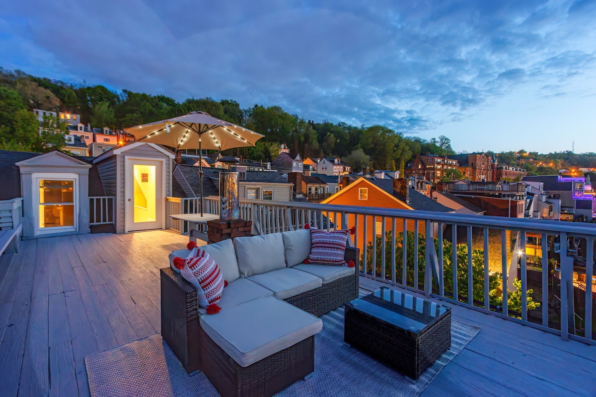 A spacious rooftop deck showcases a comfortable seating area with plush cushions and a coffee table, overlooking a vibrant cityscape. A large umbrella provides shade, while the surrounding rooftops are illuminated by soft evening light.