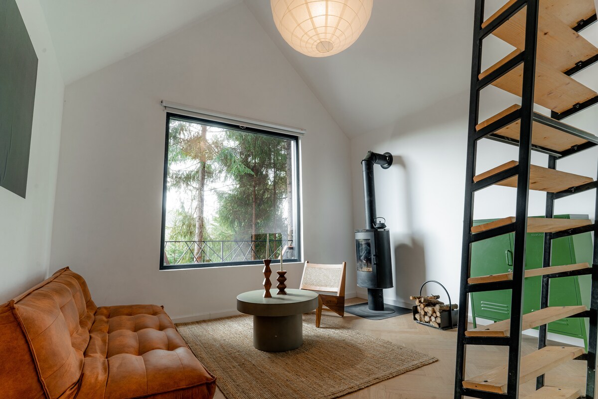 An open-plan living space features a comfortable sofa and a circular coffee table placed on a natural fiber rug. A large window offers views of surrounding trees, while a black wood stove adds warmth. A minimalist staircase provides access to the upper level.