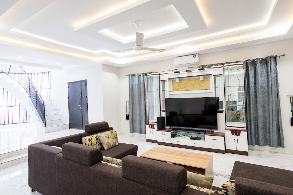 A spacious living area is distinguished by a modern design featuring a large television unit and a comfortable sectional sofa. Soft, neutral-toned walls frame the space, while large windows are adorned with elegant curtains, allowing natural light to enhance the room's inviting ambiance.