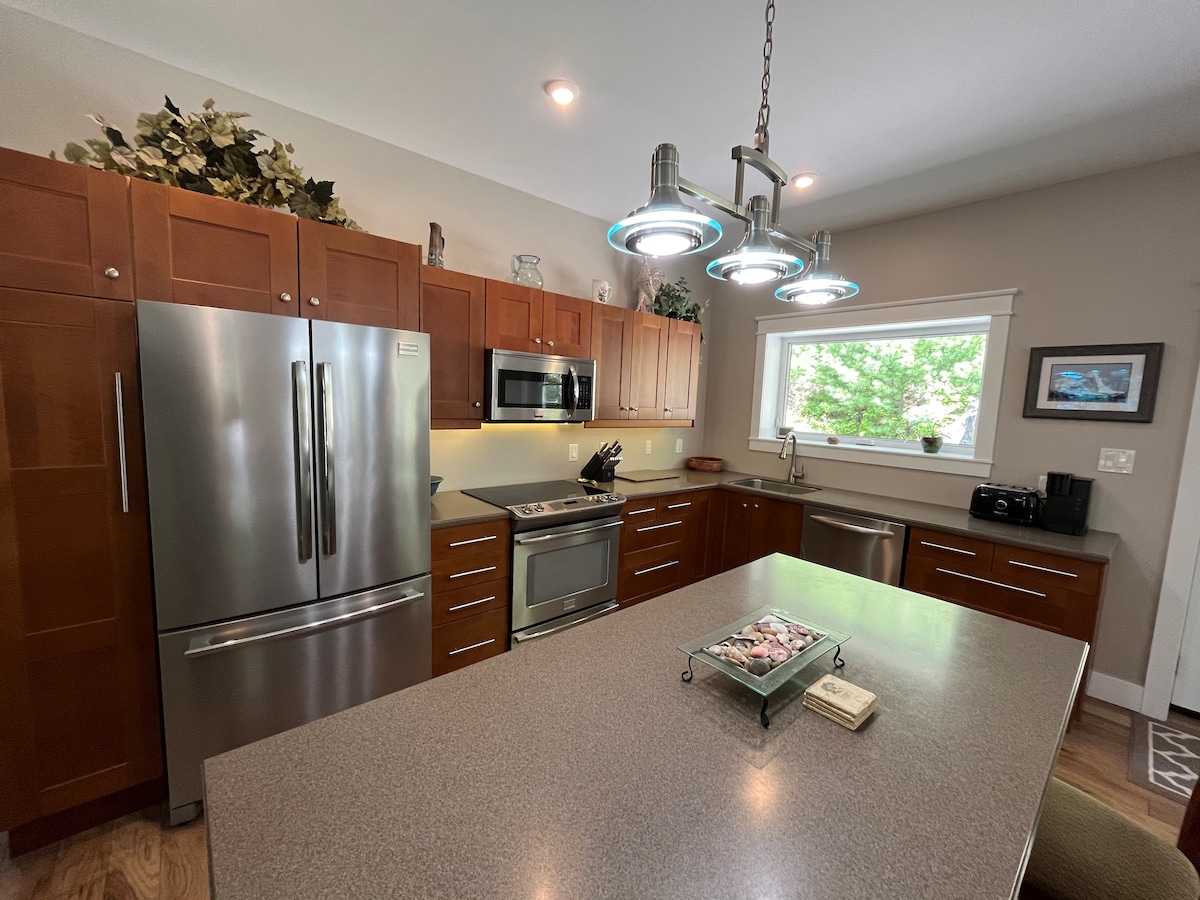 The spacious kitchen features rich wooden cabinetry and stainless steel appliances, including a refrigerator and microwave. A large island with seating is central to the layout, while a bright window provides natural light and views of greenery outside.