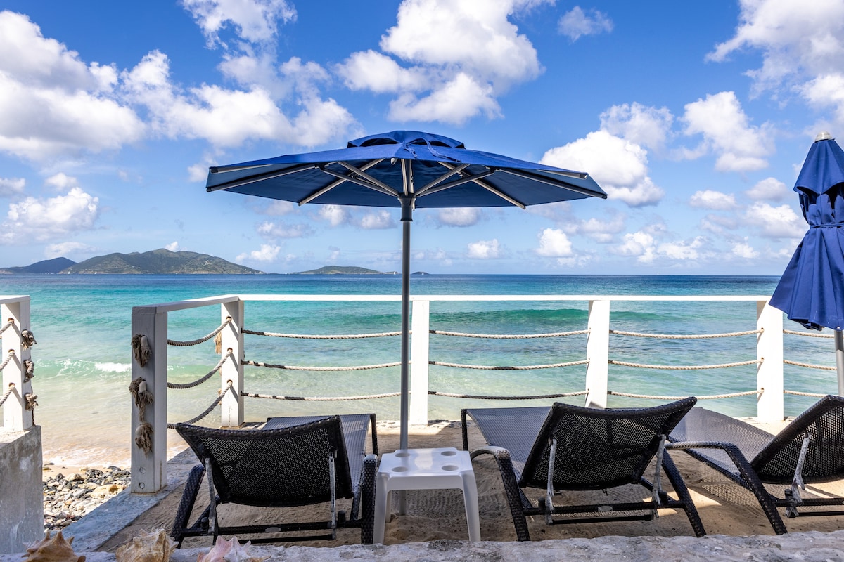 Lounge chairs are positioned by the water's edge under a blue umbrella, offering a serene view of the turquoise sea. Soft, white sand is visible in the foreground, while a few clouds drift across a clear sky in the background.