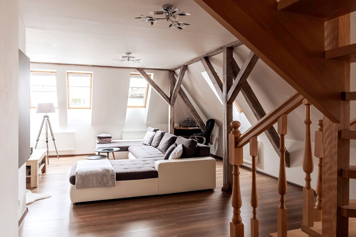 The living room showcases a spacious layout with large windows allowing natural light to fill the area. A sectional sofa is positioned on a wooden floor, accompanied by a coffee table. Distinctive wooden beams are visible, adding to the architectural character of the space.