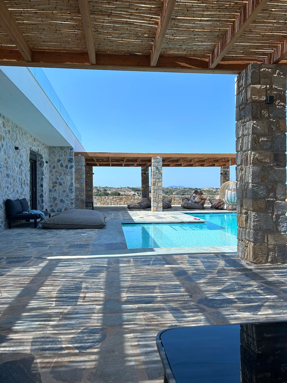 The outdoor space features a stone-paved area adjacent to a private pool. Sunlight dapples the floor through a wooden pergola overhead. In the background, comfortable seating and decorative cushions provide a relaxing retreat. The surrounding landscape and blue sky are visible beyond the terrace.