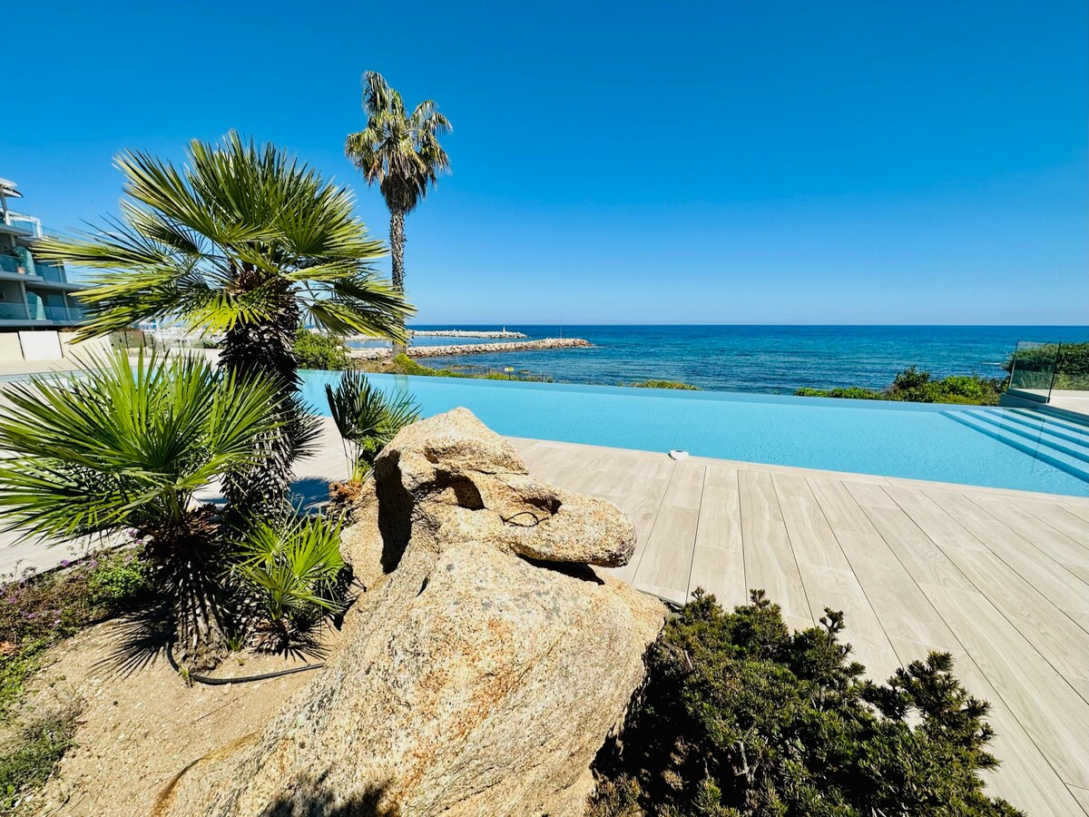 A tranquil infinity pool extends towards the ocean, surrounded by a wooden deck. Lush palm trees and rocky landscaping frame the scene, with clear blue skies reflecting over the serene waters.