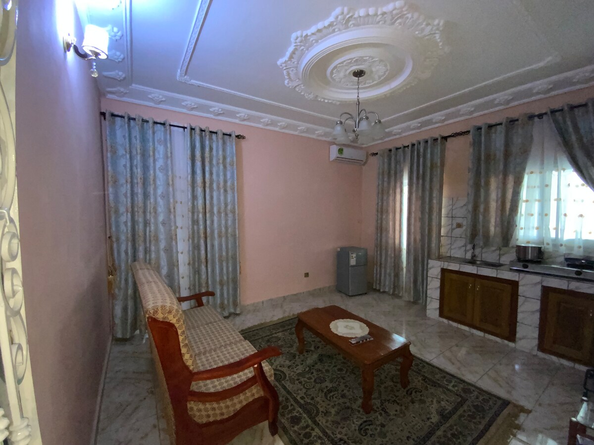 The living room features soft pink walls and elegant crown molding. A wooden coffee table sits in the center, surrounded by a patterned rug. A comfortable couch is positioned against the wall, while decorative curtains frame the windows and allow natural light to filter through.