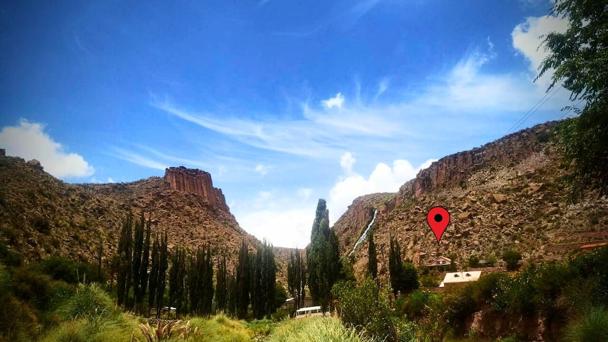 A serene landscape is shown with dramatic cliffs and vibrant greenery. Towering trees line the foreground, while a distant cabin is nestled against the mountainside. Bright blue skies with wispy clouds create a peaceful atmosphere, highlighting the natural beauty of the surroundings.