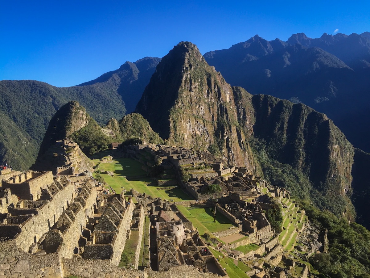 An aerial view of Machu Picchu reveals its intricate stone structures nestled among rugged mountains. Lush greenery surrounds the site, with terraced fields and walkways visible in the foreground, leading to the iconic peaks in the background against a clear blue sky.
