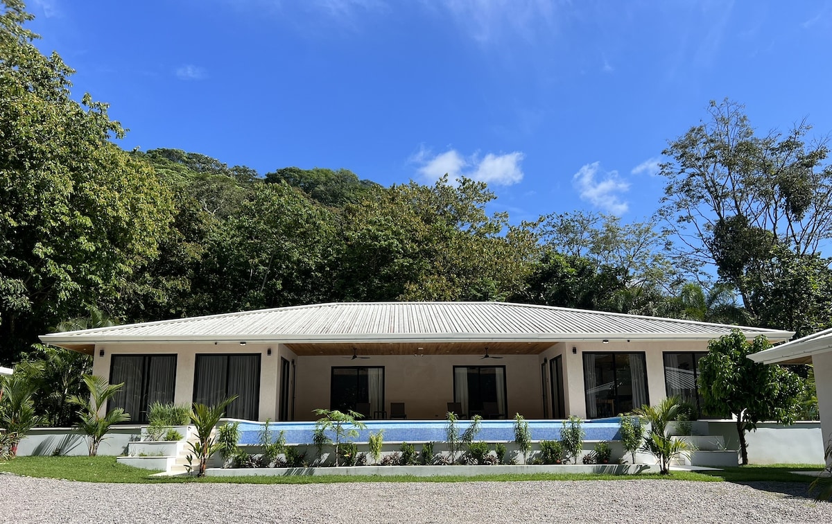 The exterior of a modern villa is highlighted by a sleek architecture and large windows. Lush greenery surrounds the property, with an inviting infinity pool visible in front. A gravel pathway leads to the entrance, enhancing the serene atmosphere.