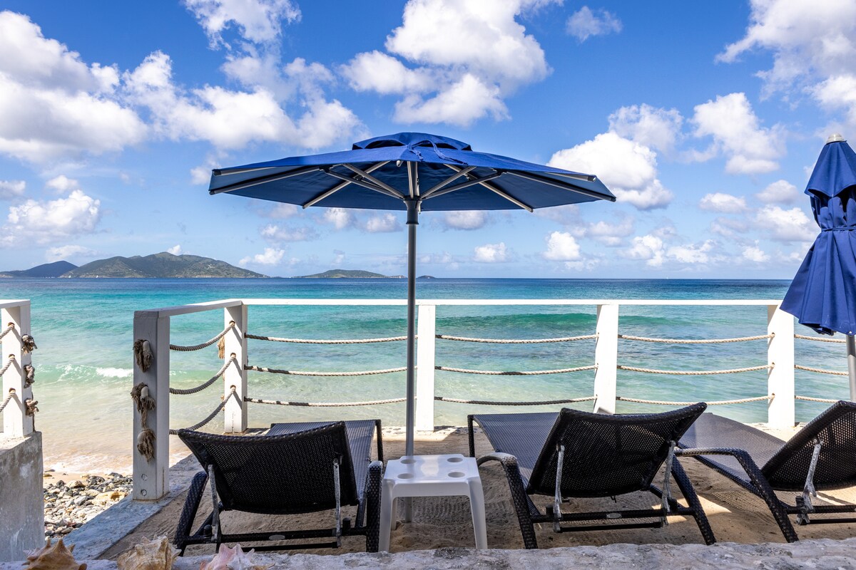 A serene view is offered from the beachside loungers, shaded by a large blue umbrella. Soft waves lap against the shore, with distant islands visible on the horizon. The sky is partly cloudy, enhancing the tranquil atmosphere of the oceanfront setting.