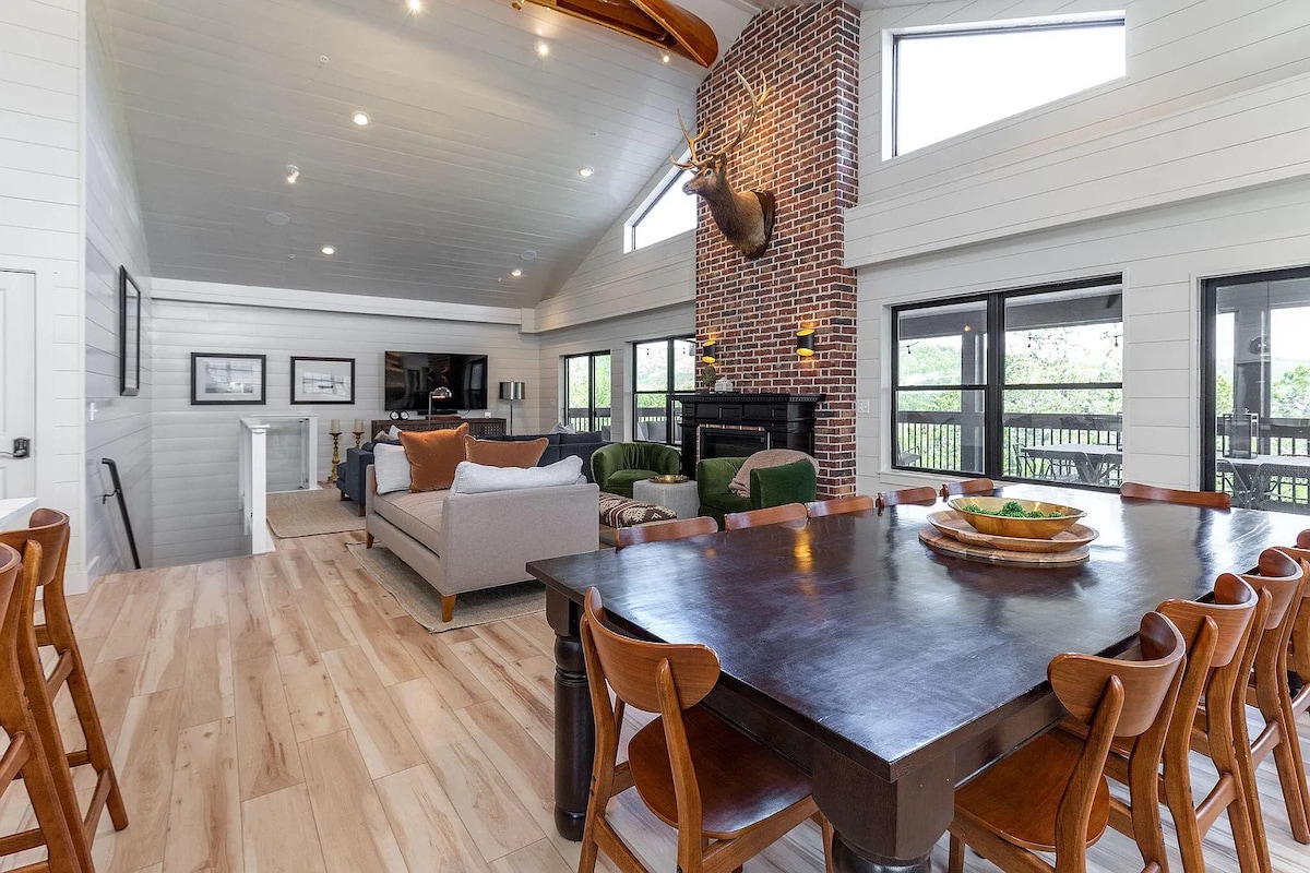 An open concept living area features a large brick fireplace and high vaulted ceilings. A dark wooden dining table with chairs is positioned at the forefront. Comfortable seating, including a light-colored sofa, is arranged to foster socializing, with large windows offering views of the surroundings.
