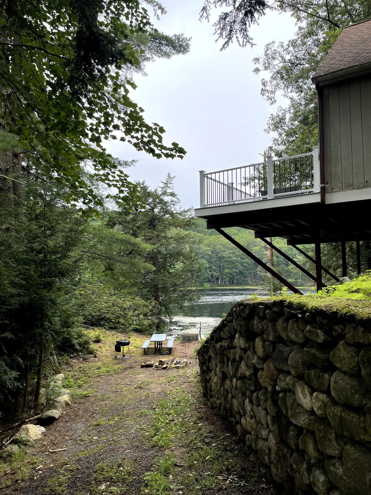 A path lined with lush greenery leads toward a stone wall. A shaded area is visible, featuring a grill and picnic table, with a view of the serene lake in the background. The overhanging deck of a house is seen above the path.