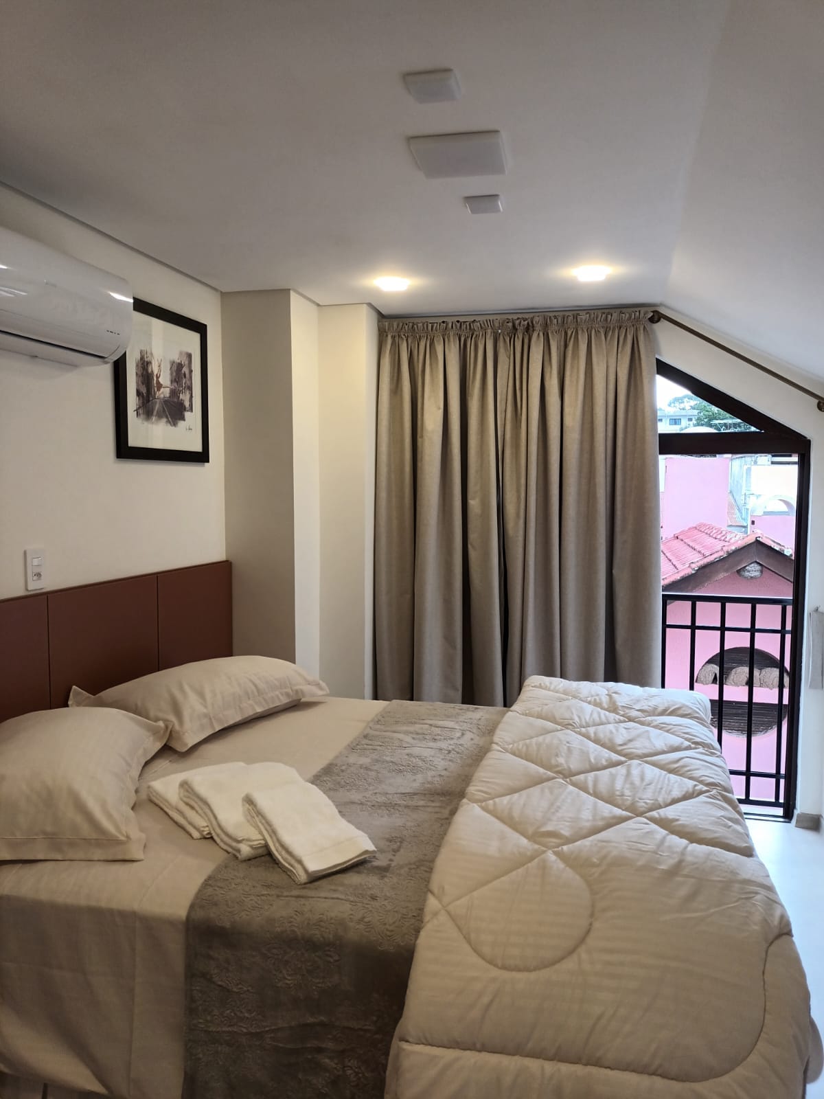 A comfortable sleeping area features a spacious bed with neutral-toned bedding and folded towels. Natural light enters through a window, and soft curtains frame the view. An air conditioning unit is installed above the bed, and a framed picture adds a touch of decor.