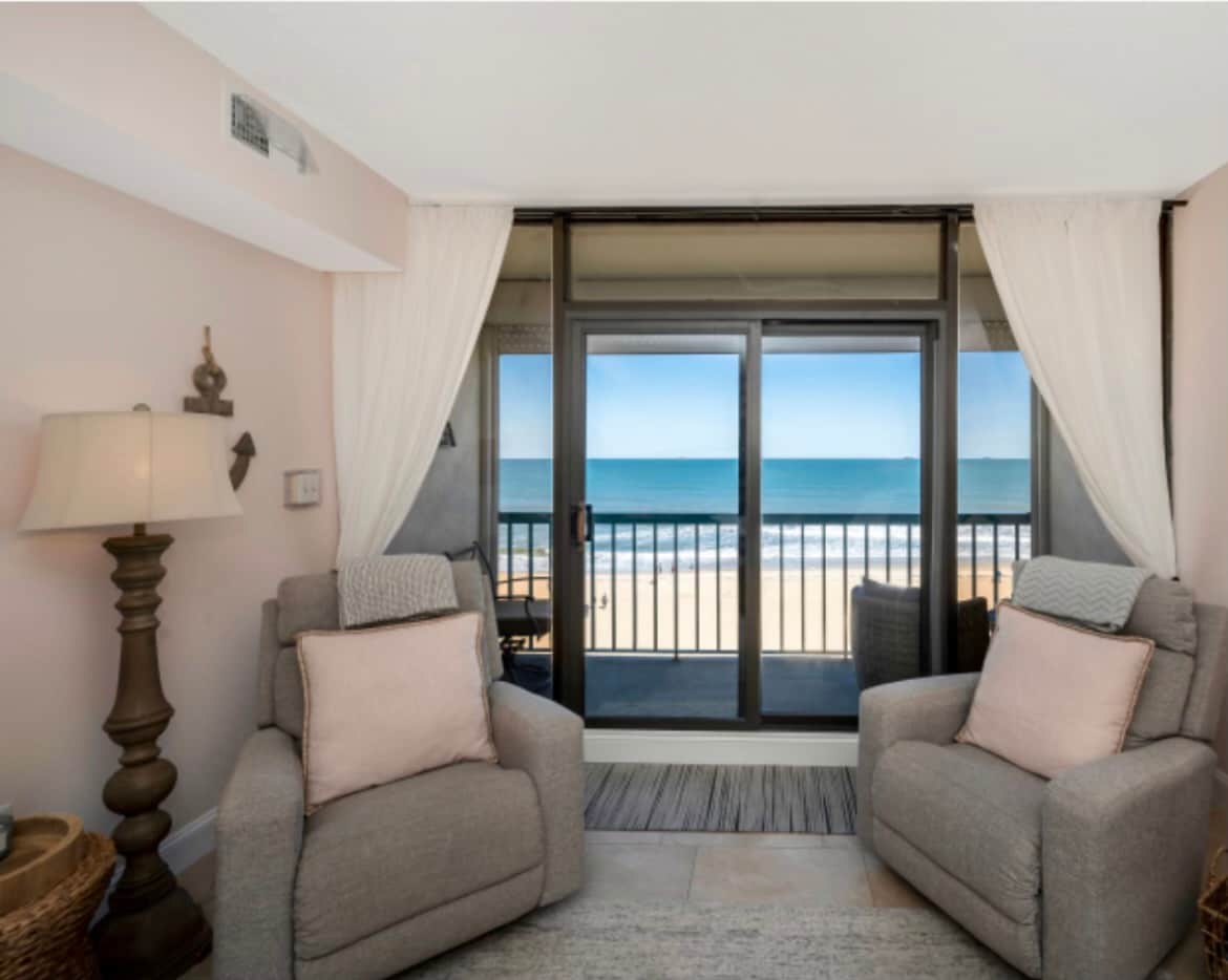 Two comfortable chairs are positioned in front of large sliding glass doors, providing an unobstructed ocean view. Soft curtains frame the windows, and a decorative table lamp adds warmth to the inviting space. The room features a neutral color palette with subtle coastal accents.