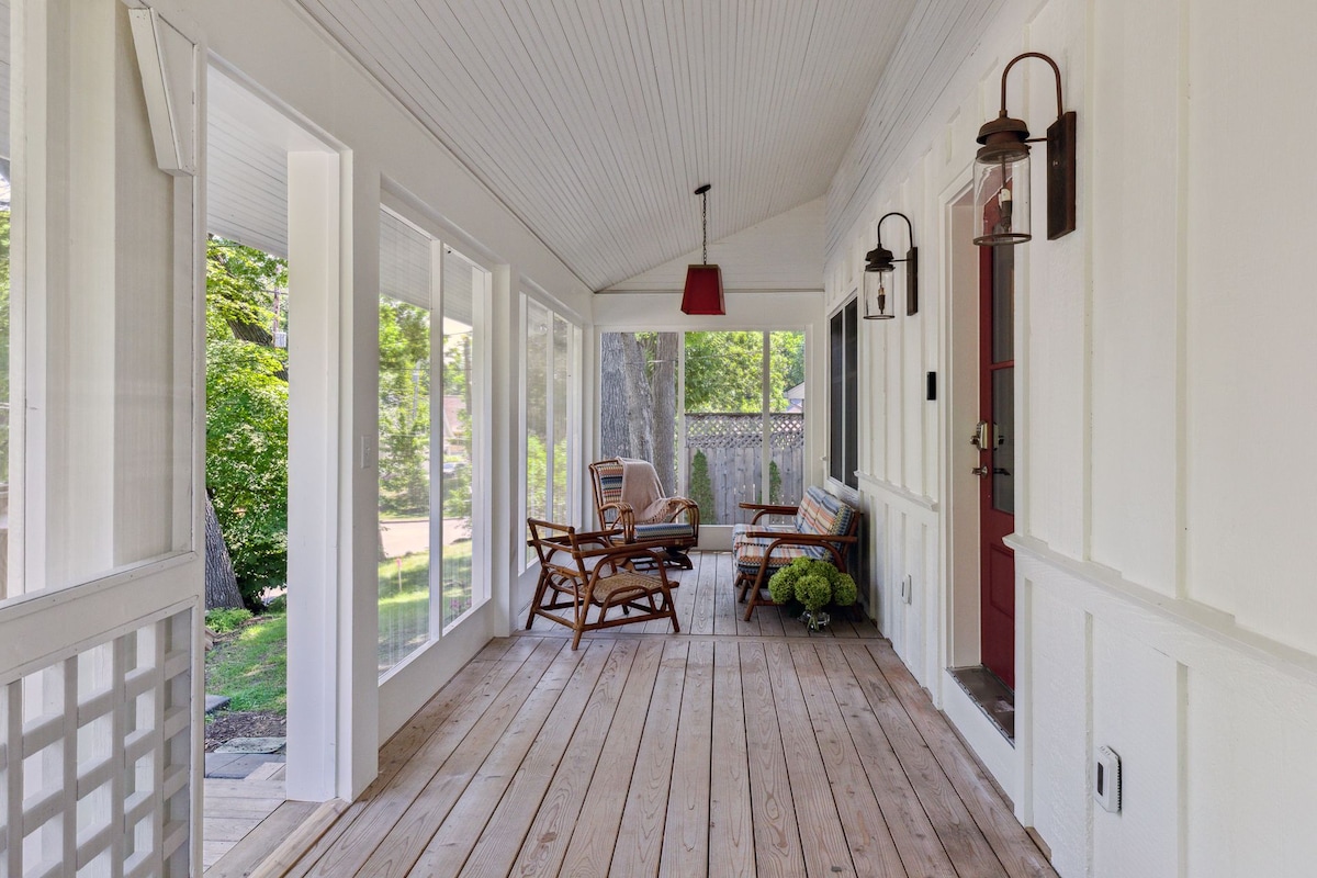 A screened-in porch features wooden flooring and white walls, providing a spacious area for relaxation. Comfortable seating arrangements include two rocking chairs and a small table. Natural light floods the space through large windows, with greenery visible outside.