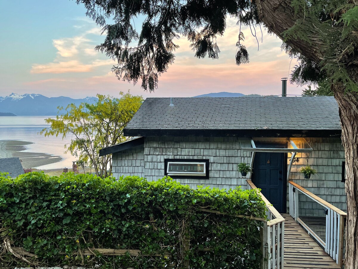 The exterior of 'The Coast House' is shown, framed by lush greenery. A wooden footbridge leads to the entrance, highlighting a welcoming blue door. In the background, the serene waterfront reflects soft pastel colors of a sunset, with distant mountains visible beyond.