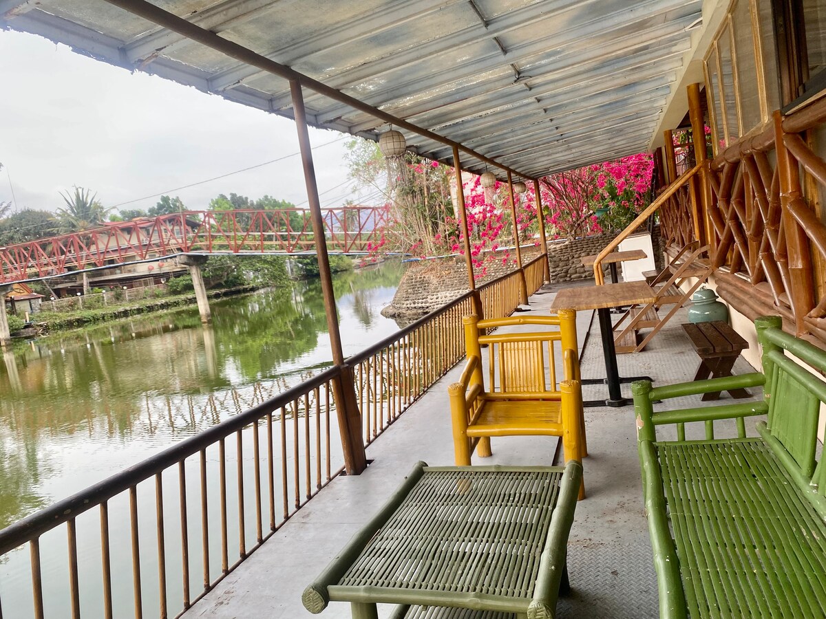 A covered porch features a combination of yellow and green seating options, with a low table positioned between them. Lush pink bougainvillea drapes over the railing, while a calm waterway reflects the surroundings. A red bridge can be seen nearby, enhancing the scenic setting.