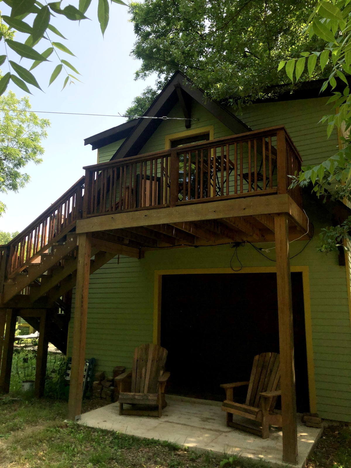 A two-story structure features a wooden deck with seating for relaxation, accessed by external stairs. The siding is a soft green, surrounded by trees. Two sturdy wooden chairs sit on a concrete patio beneath the deck, inviting guests to enjoy the outdoor space.