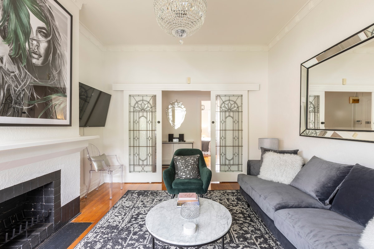 The living room is highlighted by a plush velvet sofa and accent chairs, paired with an elegant coffee table. A statement crystal chandelier hangs above, while the large mirror enhances the room's spaciousness. Decorative glass doors lead to an adjoining area, adding charm and sophistication.