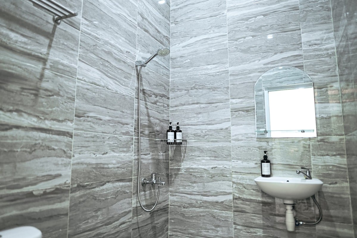 A modern bathroom is shown with sleek grey marble tiles covering the walls. A shower with a glass divider features an overhead rain showerhead. A mirror is mounted above a small sink, accompanied by stylish toiletries on a wall-mounted shelf.