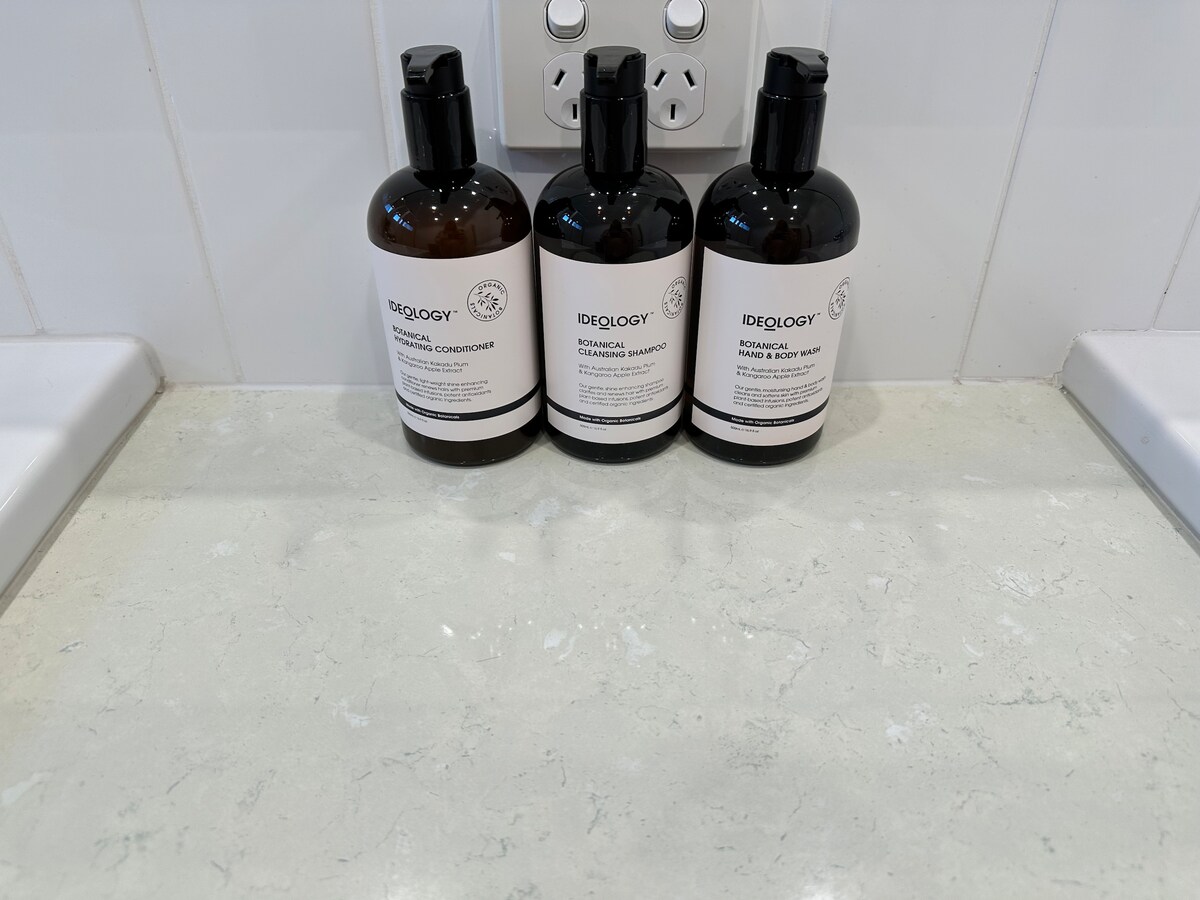 Three bottles of shampoo, conditioner, and body wash are neatly arranged on a light-colored countertop. Each bottle displays a clear label with product details, set against a backdrop of white tiles and visible electrical outlets.