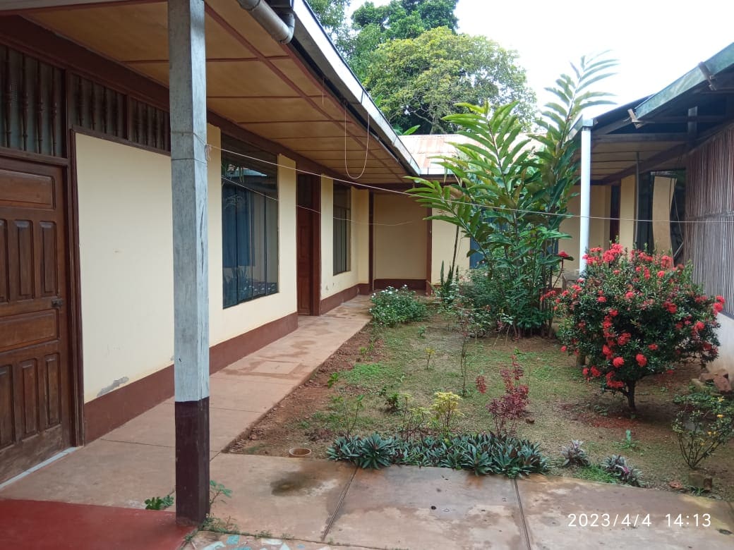 A serene courtyard is surrounded by a mix of one-story buildings. Lush greenery and flowering plants adorn the garden, providing a natural touch. Pathways lead through the area, with wooden doors visible on one side, contributing to the tranquil atmosphere.