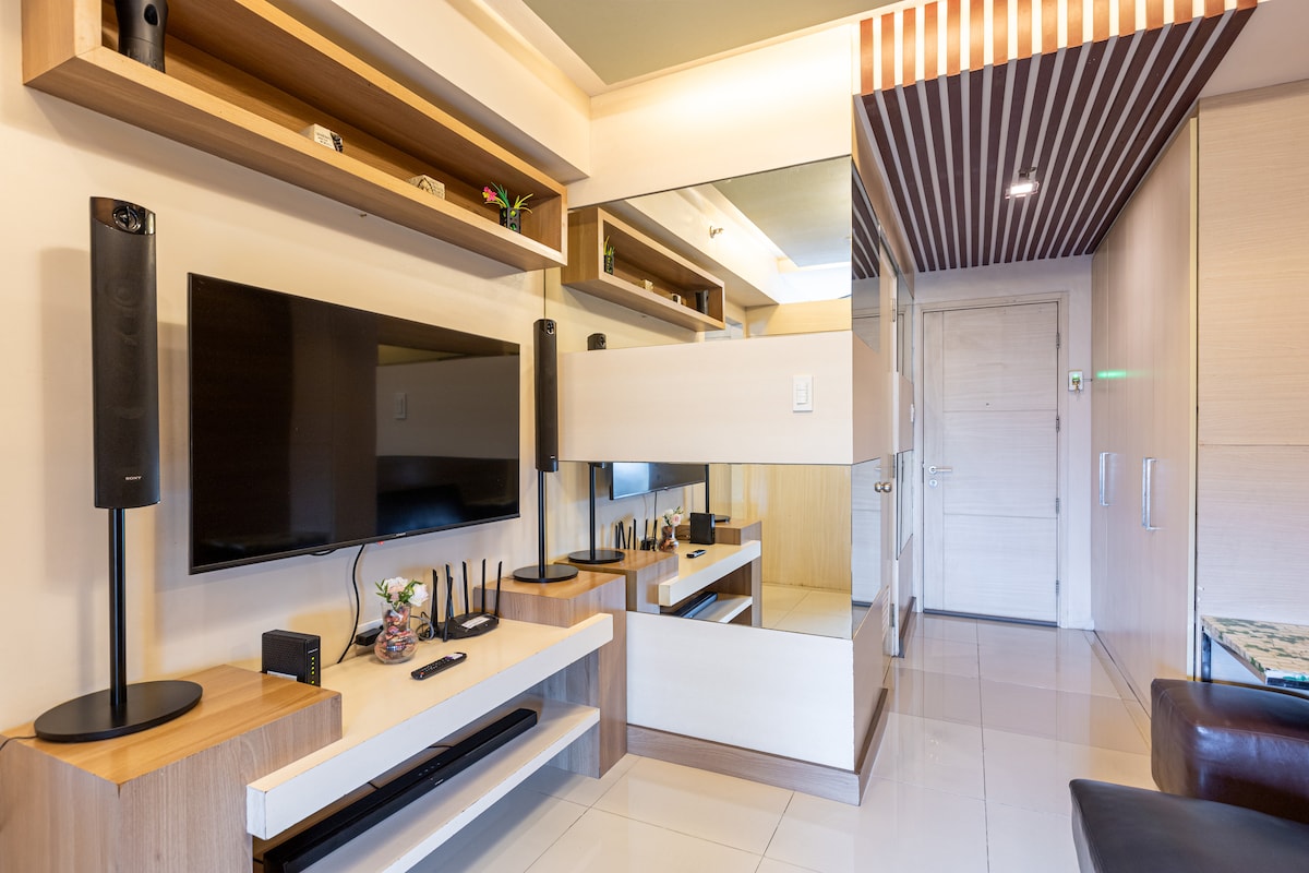 A modern living area is presented, featuring wall-mounted shelves and a large flat-screen TV. A sleek mirror reflects the room's layout, while a leather couch adds seating comfort. Decorative accents and a sound system complement the space, creating a functional entertainment area.