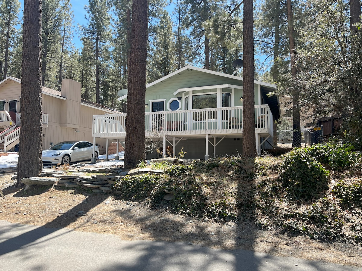 A charming single-story home is situated among tall pine trees. A spacious front deck is visible, surrounded by natural landscaping. A car is parked in the driveway, and the structure features a rounded window that allows light into the interior.