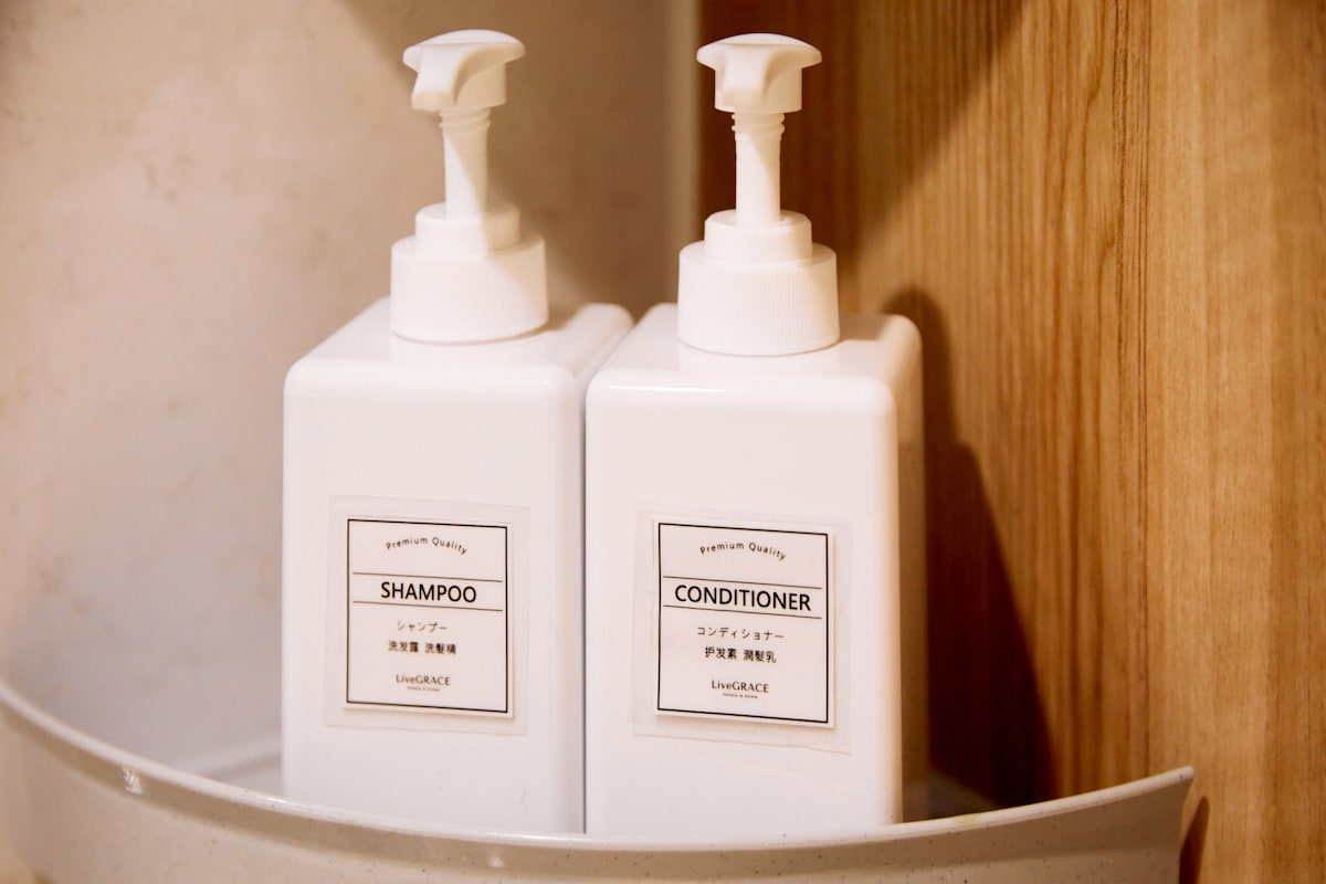 Two white bottles labeled 'SHAMPOO' and 'CONDITIONER' are neatly placed on a light-colored shelf. Each bottle features a pump dispenser, providing easy access to the toiletries. The warm wooden background adds a natural touch to the bathroom space.