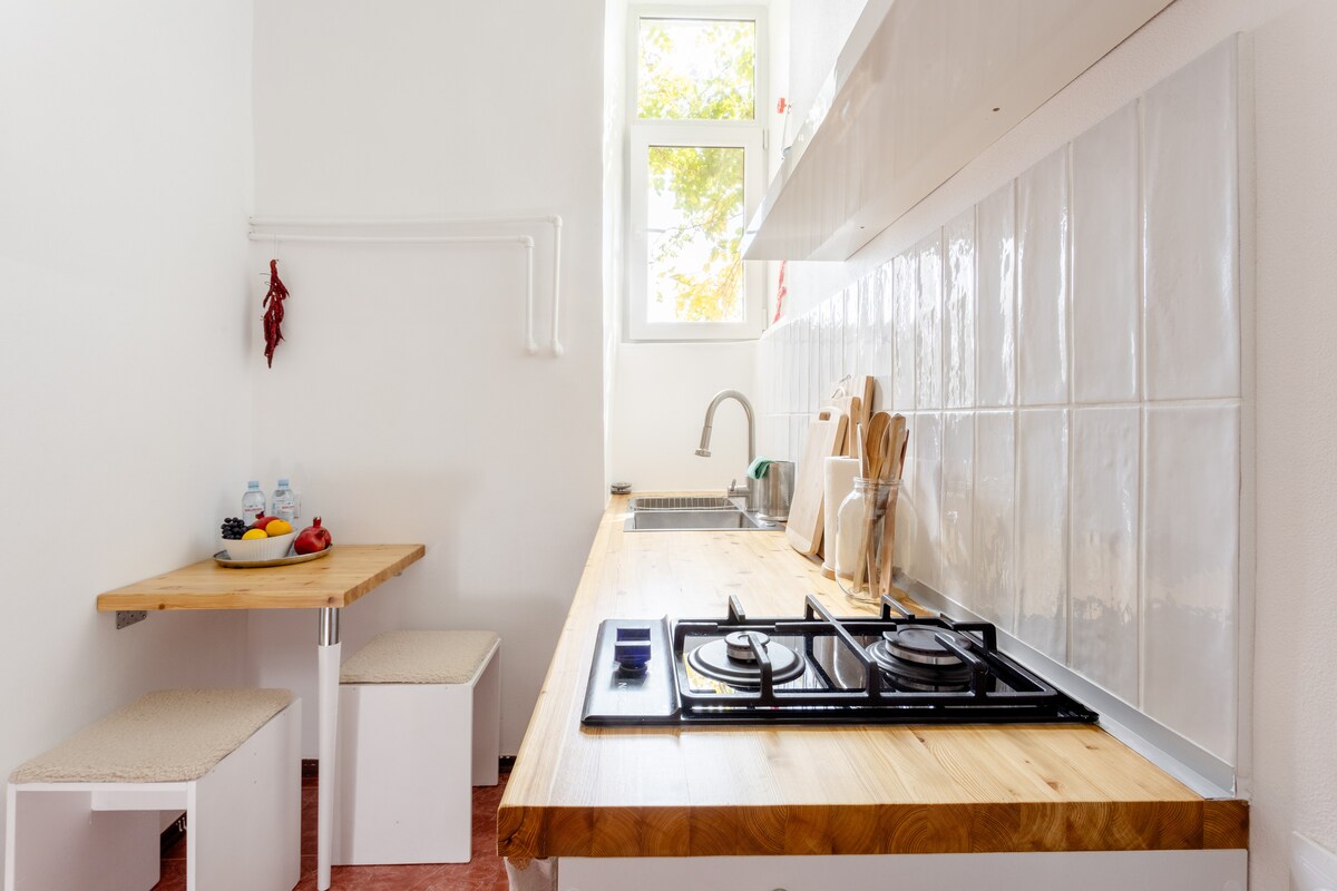 A bright kitchen space features a wooden countertop with a gas stove and organized utensils. A small table with two stools is visible, accompanied by a bowl of fresh fruit. Natural light flows through the window, illuminating the clean, white tile backsplash.