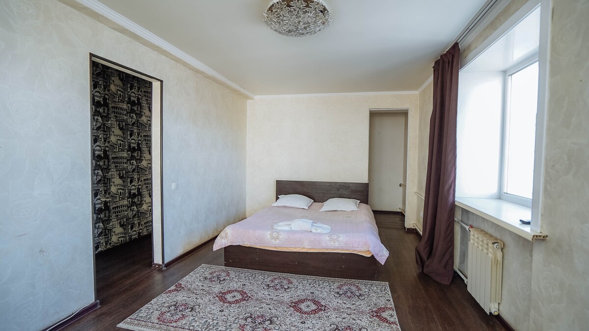 A spacious bedroom features a large bed with light bedding, flanked by two side tables. Natural light floods the room through a wide window, while a decorative area rug lays on the polished floor. A muted wallpaper adds subtle texture to one wall.