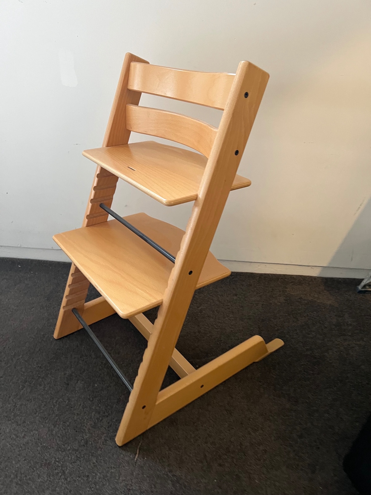 A wooden high chair is presented in a light finish, showcasing three adjustable seating levels. The chair features a sturdy design with a wide base for stability and has a simple, modern aesthetic suitable for various settings.