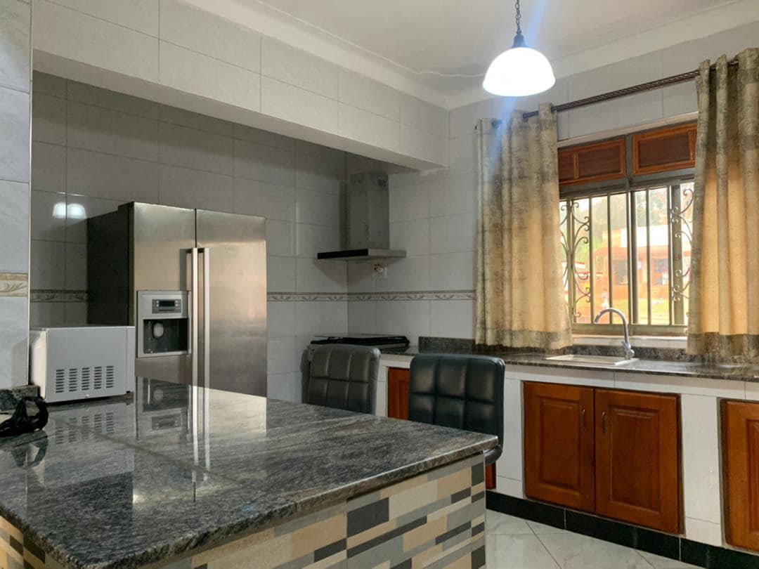 The kitchen features a granite countertop with a breakfast bar and dark wooden cabinets. Stainless steel appliances, including a refrigerator and built-in microwave, are visible. A window provides natural light, framed by curtains, while the tiled floor offers a clean and contemporary look.