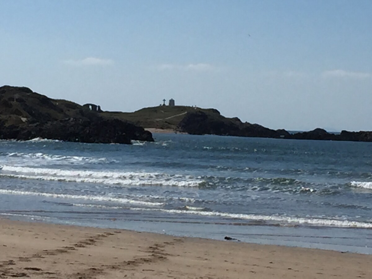 The image showcases a sandy beach with gentle waves lapping at the shore. In the distance, an island is visible, featuring structures that could be a lighthouse or similar buildings. Clear blue skies with a few clouds complete the serene coastal scene.
