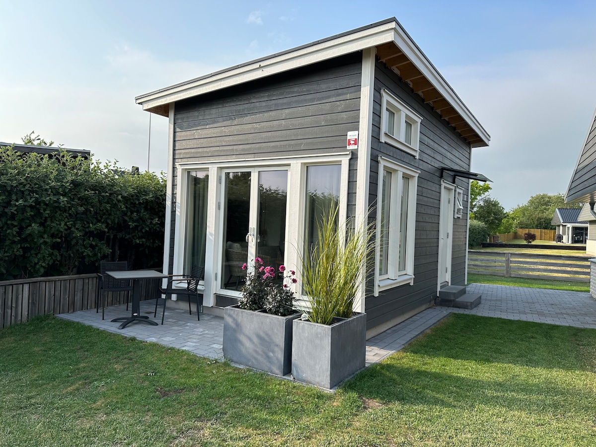 A modern Attefall house is depicted with large glass doors, allowing natural light to fill the interior. A small patio area features black outdoor furniture beside planters containing vibrant flowers. The surrounding green lawn and hedges create a peaceful outdoor setting.