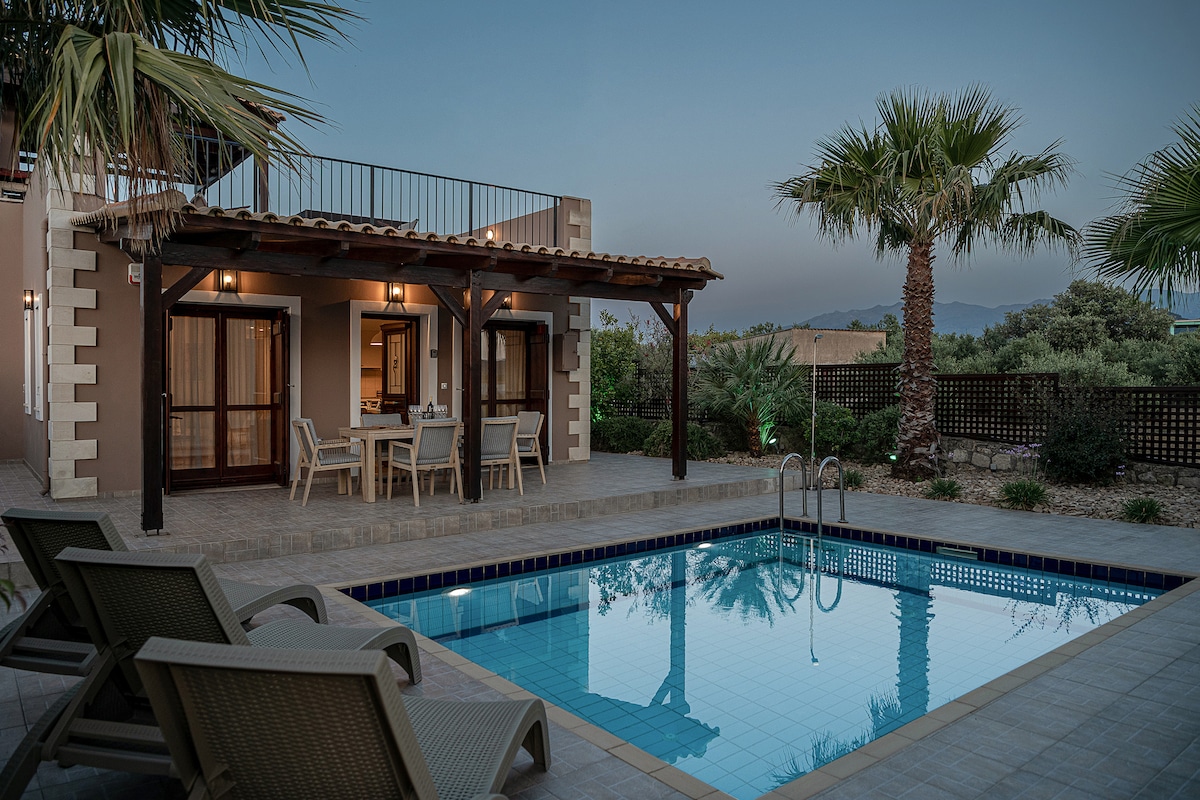 A private pool area is visible, featuring a rectangular swimming pool surrounded by tiled flooring. Four lounge chairs are positioned beside the water, offering a place to relax. The villa's exterior is characterized by wooden beams and large glass doors, enhancing the connection with the outdoor scenery.