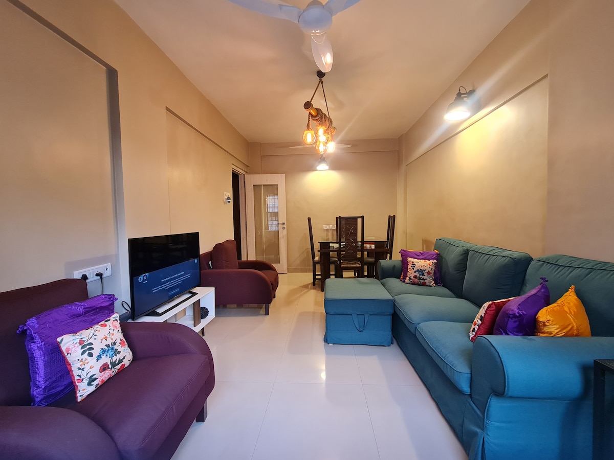 A spacious living area features a combination of comfortable seating with a deep blue sofa and purple armchairs adorned with colorful cushions. A dining table is positioned under elegant lighting, complemented by soft wall colors and a television mounted on one wall.