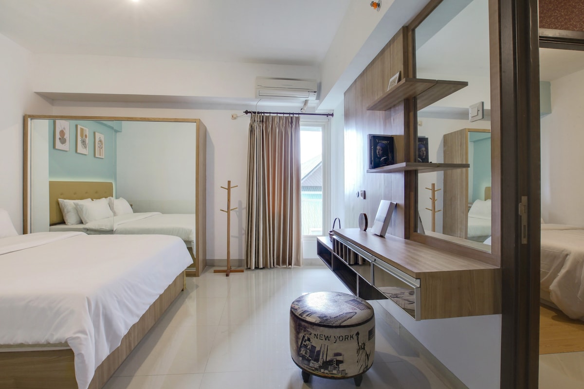 A bright and spacious bedroom features two distinct sleeping areas with soft bedding. A large mirror reflects the room's light and modern decor. Natural light filters through the window, complemented by smooth, neutral-toned walls and contemporary furnishings.