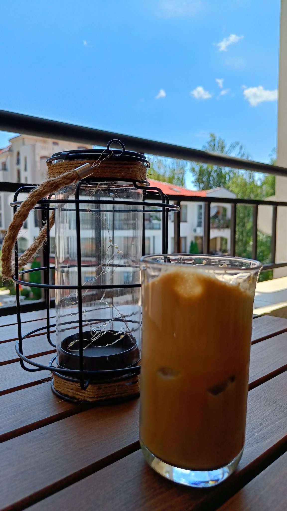 A refreshing iced coffee is shown in a glass with ice cubes, placed on a wooden table. A decorative lantern, featuring a glass body and black wire frame, sits nearby, with clear blue skies and greenery visible in the background.