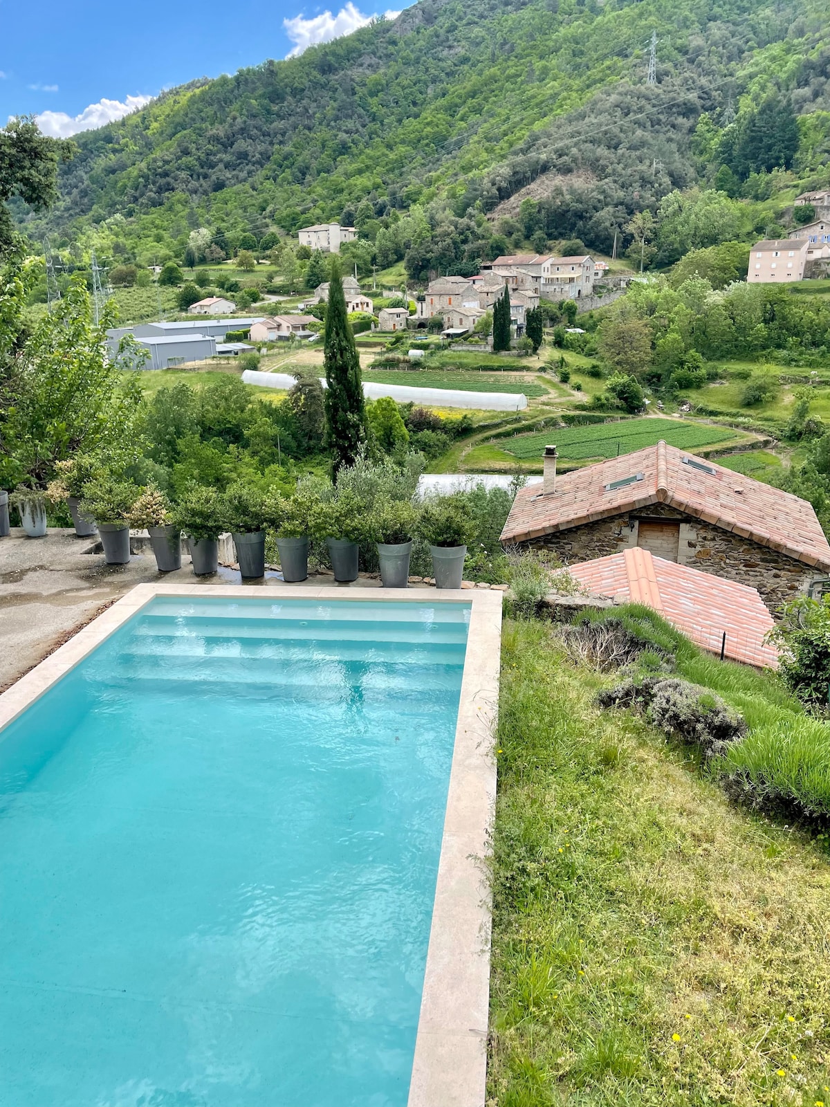 A salt-water pool is seen nestled within a serene landscape, surrounded by greenery. The hillside is dotted with quaint houses and fields, while mountains provide a scenic backdrop, enhancing the natural beauty of the area.