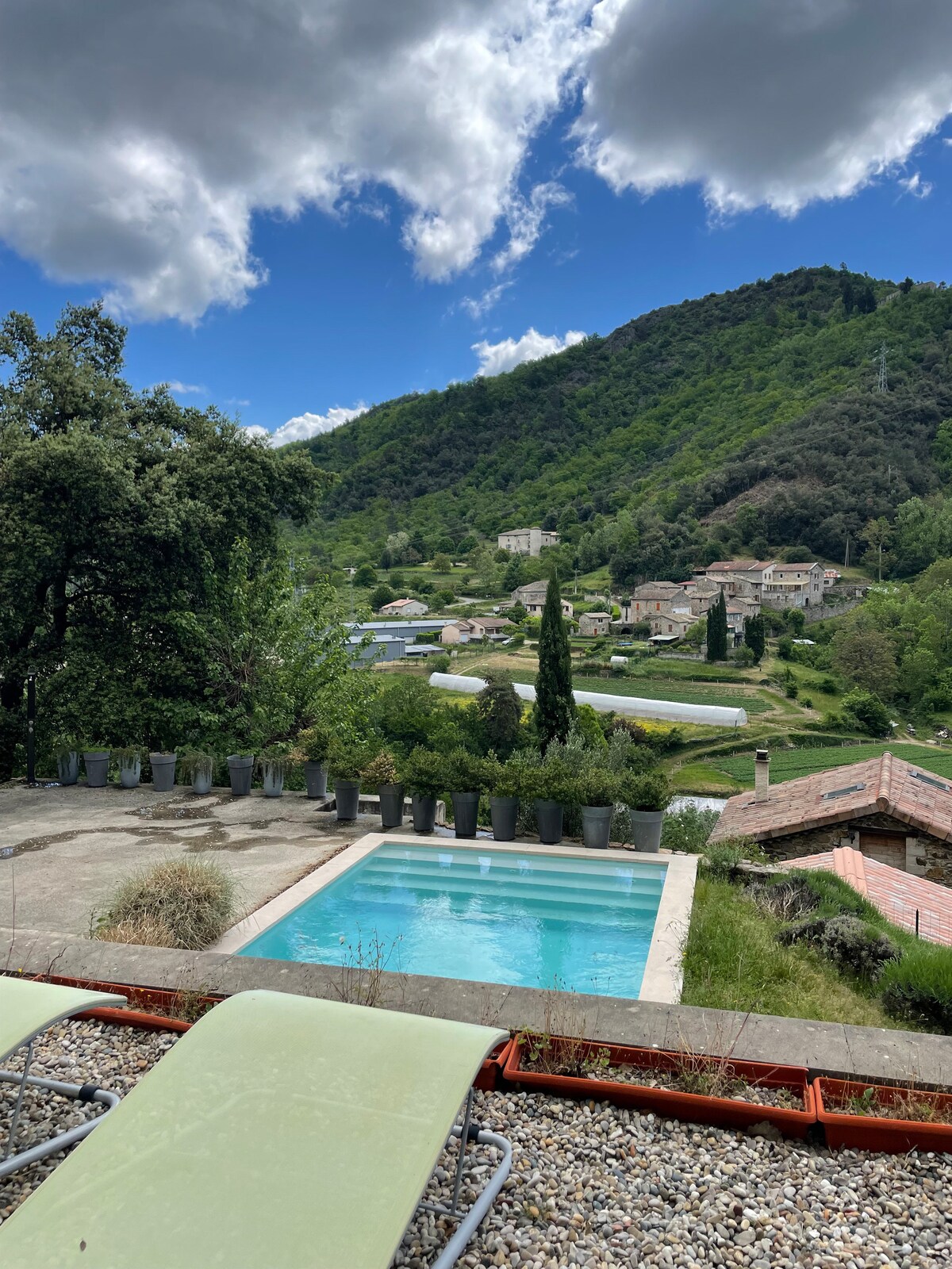 A serene view showcases a salt-water pool bordered by concrete and potted plants, with a backdrop of lush green hills and the village below. The sky is partly cloudy, adding depth to the landscape. Sun loungers are positioned on the terrace in the foreground.