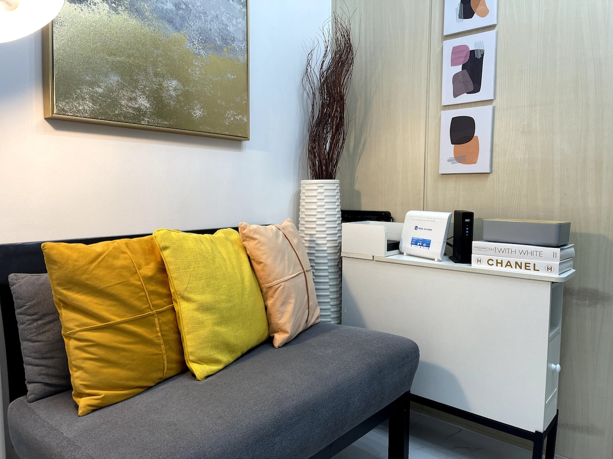 A cozy living area is presented with a two-seater sofa adorned with yellow and pastel cushions. A white cabinet is positioned beside the sofa, featuring decorative items and books. A piece of abstract art hangs on the wall above, enhancing the modern aesthetic.